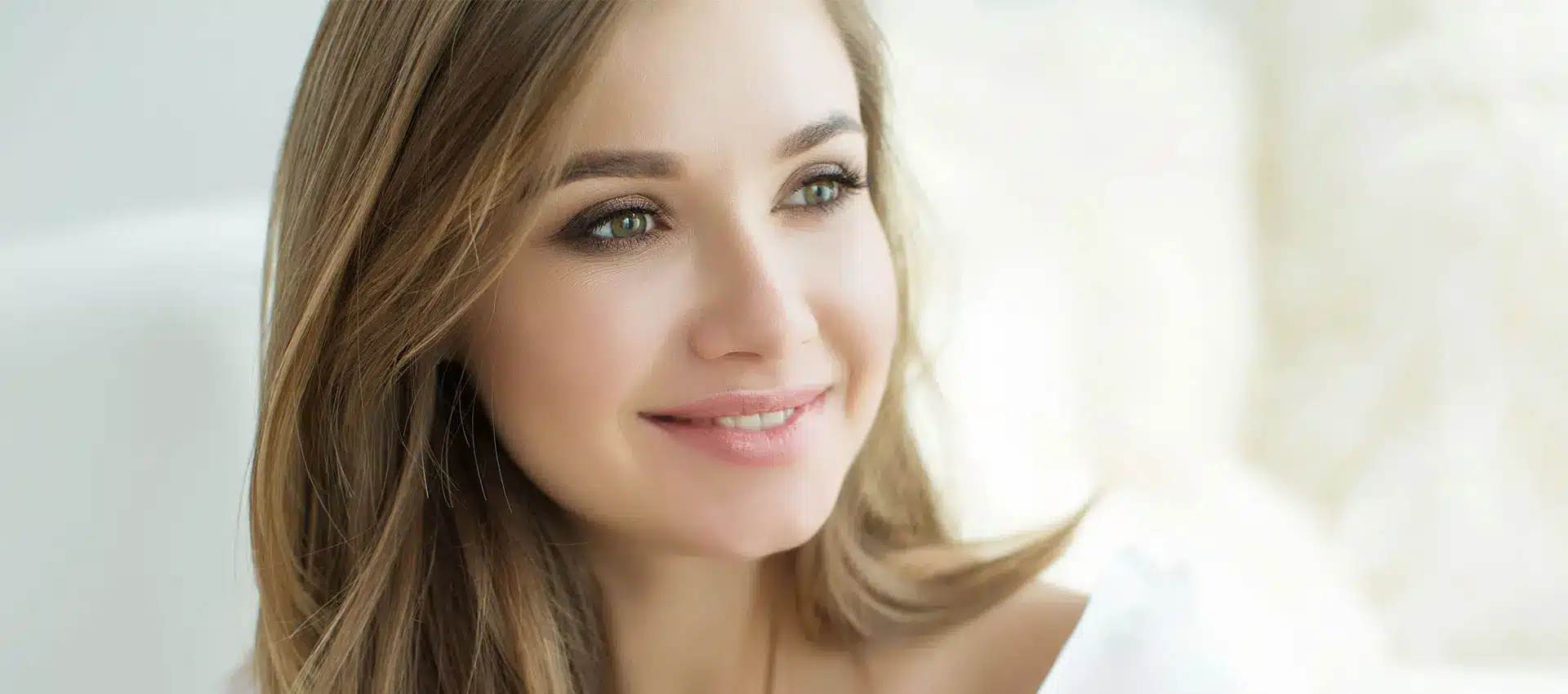 a woman with long light brown hair and green eyes is smiling softly she is wearing a white top and looking slightly to the right the background is blurred light and airy giving the image a serene and peaceful ambiance reflecting her recent cheek augmentation that enhances her natural beauty